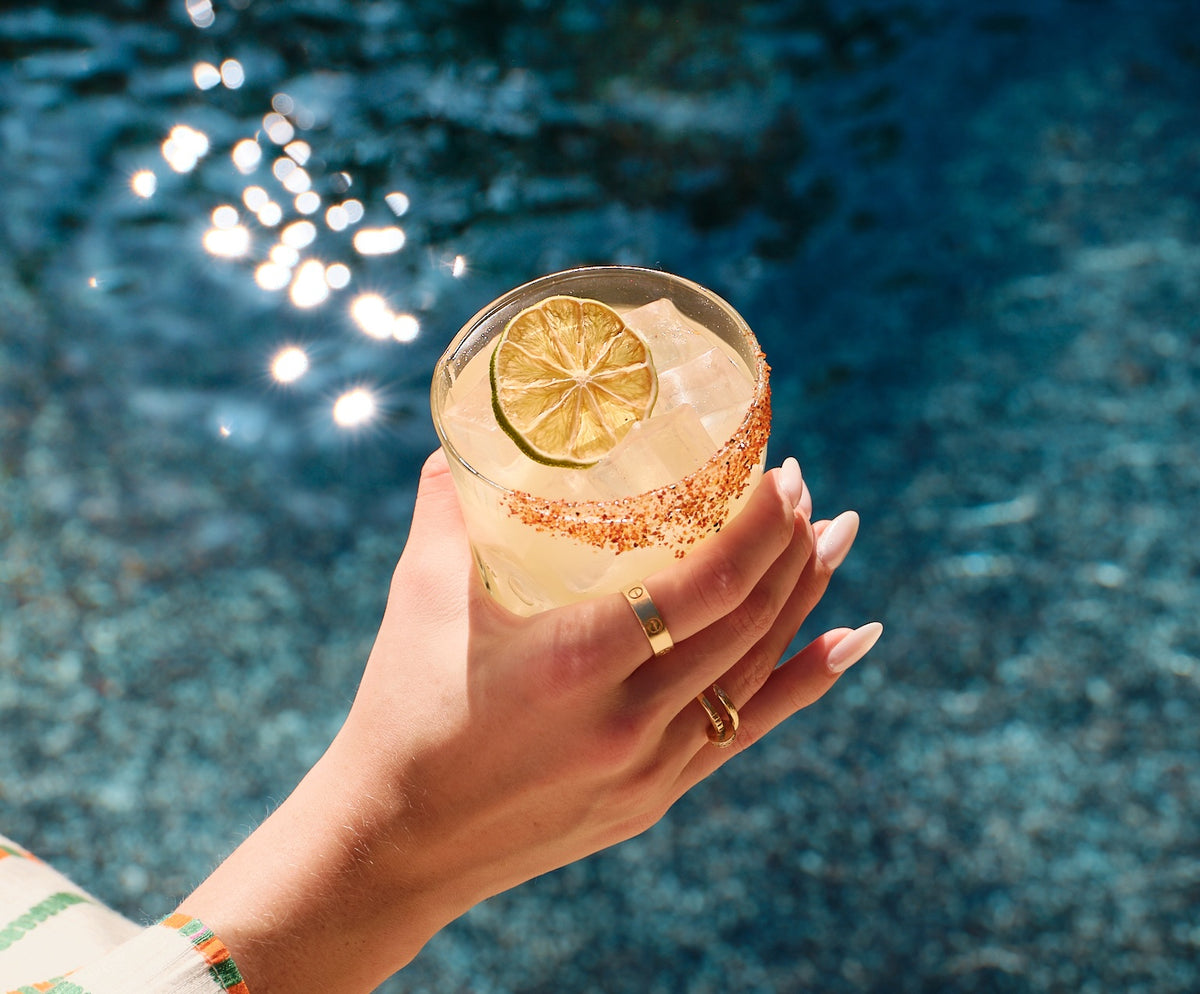 A hand holding a margarita rimmed with chili salt and garnished with a dried lime wheel, above a sunlit pool