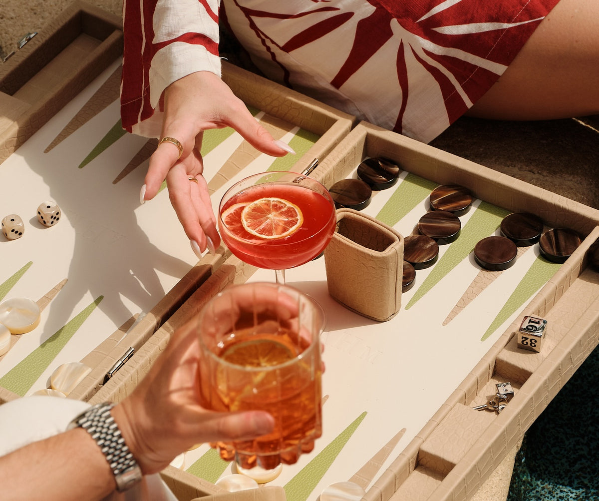 Two cocktails garnished with dried orange wheels, held over a backgammon board in afternoon sun